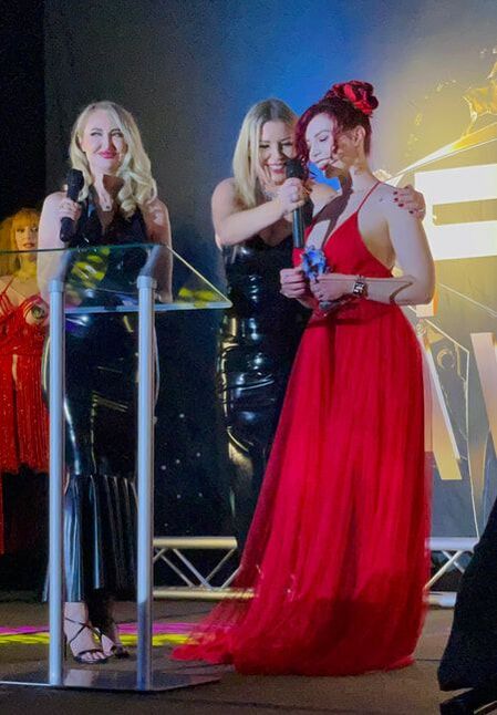 Ludella Hahn accepting one of seven awards from hosts Anastasia Pierce and Kendra James at the 2022 Fetish Awards